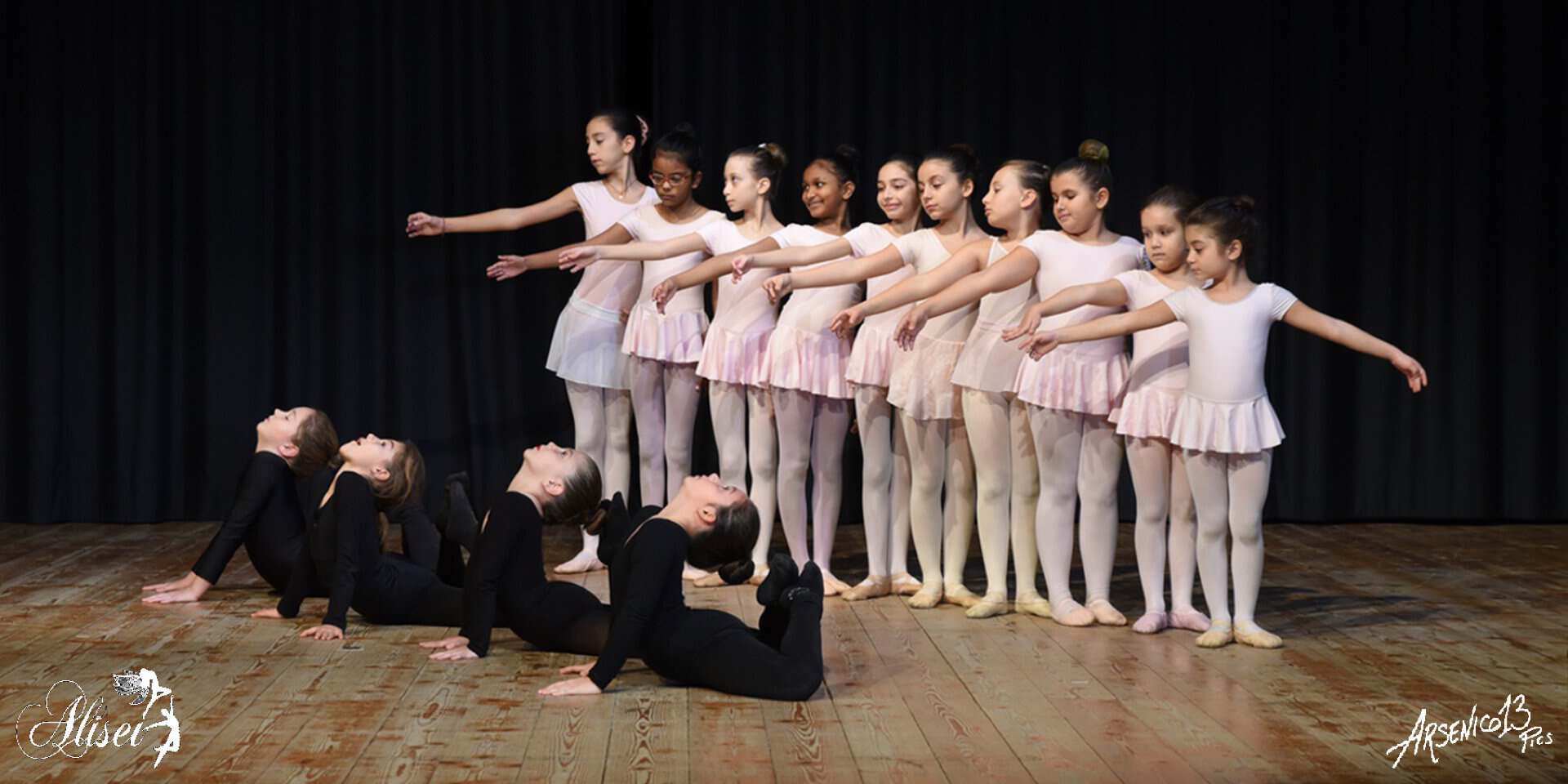 Ballet dancer in white tutu performs en pointe center stage with ensemble in blue lighting and stage lights.
